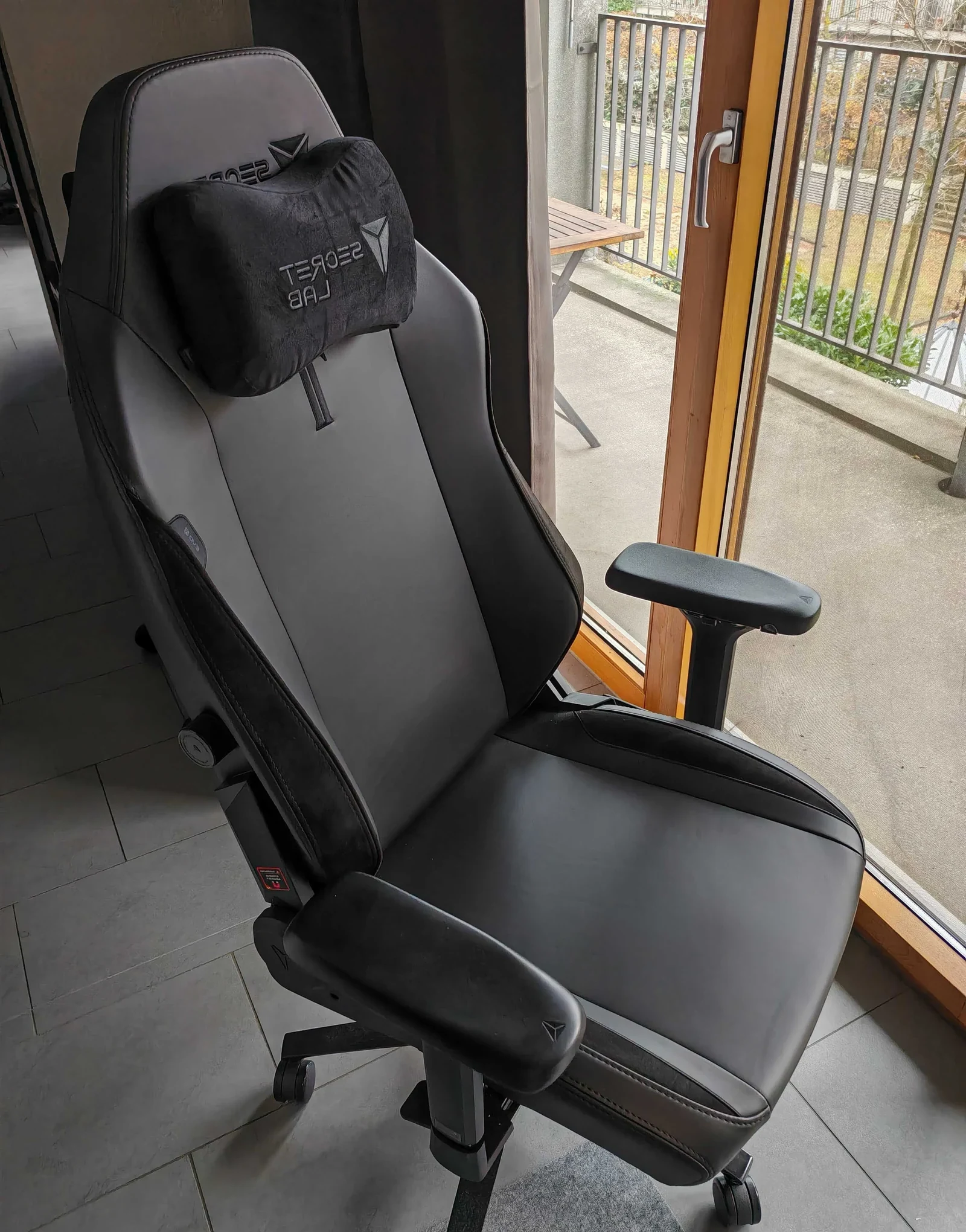 Real hands-on photo of a black Secretlab TITAN Evo chair with headrest pillow in a home office next to a balcony door (mirrored, cropped)